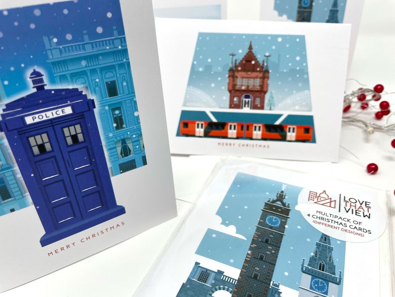 May include: A multipack of Christmas cards featuring four different designs. The cards showcase illustrations of buildings and landmarks, including a blue police box, a train, and a clock tower, with the words "MERRY CHRISTMAS" on some cards.
