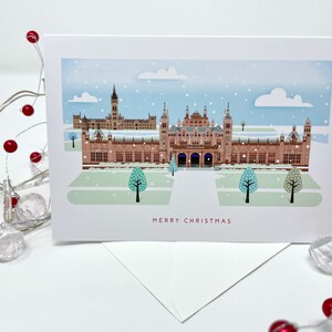 May include: A Christmas card featuring a detailed illustration of a building with multiple towers and arches, under a snowy sky. The card reads "MERRY CHRISTMAS" in red text. A white envelope is visible beneath the card, with decorative red berries and lights in the foreground.