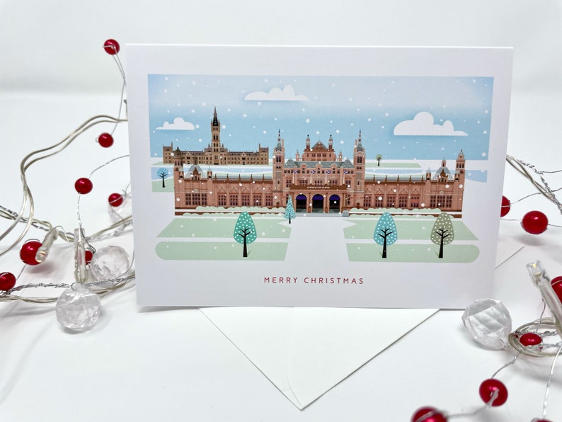 May include: A Christmas card featuring a detailed illustration of a building with multiple towers and arches, under a snowy sky. The card reads "MERRY CHRISTMAS" in red text. A white envelope is visible beneath the card, with decorative red berries and lights in the foreground.