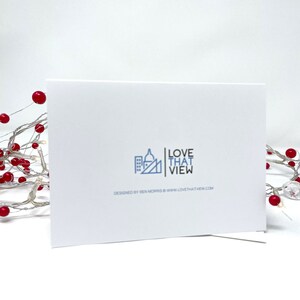May include: A white greeting card with the text "LOVE THAT VIEW" and a cityscape logo. The card is propped up against a white background. The words "DESIGNED BY BEN MORRIS WWW.LOVETHATVIEW.COM" are printed below the logo. A string of lights with red berries is in the background.
