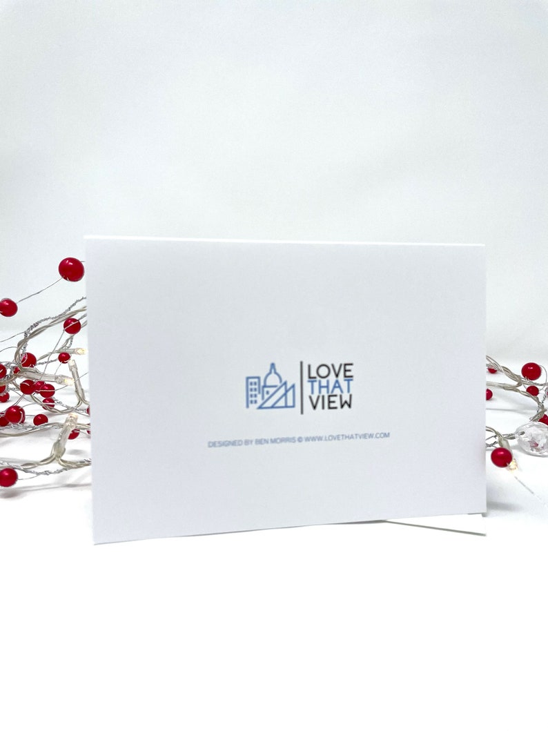 May include: A white greeting card with the text "LOVE THAT VIEW" and a cityscape logo. The card is propped up against a white background. The words "DESIGNED BY BEN MORRIS WWW.LOVETHATVIEW.COM" are printed below the logo. A string of lights with red berries is in the background.