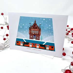 May include: A white greeting card with a winter scene featuring a red and orange train, a brick building, and falling snow. The words "MERRY CHRISTMAS" are printed at the bottom of the card.