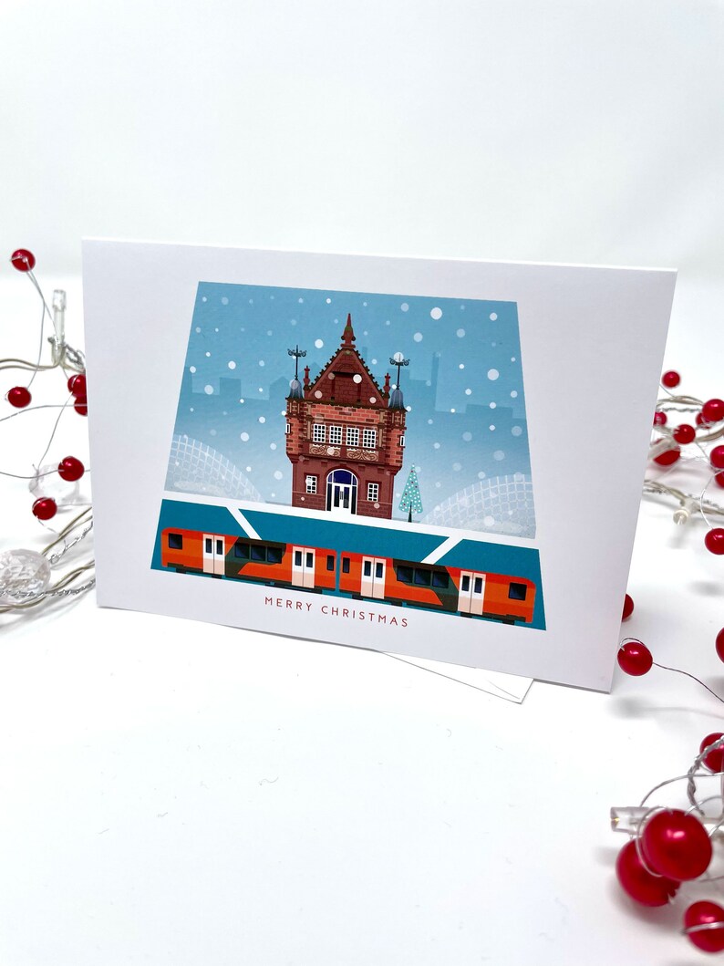 May include: A white greeting card with a winter scene featuring a red and orange train, a brick building, and falling snow. The words "MERRY CHRISTMAS" are printed at the bottom of the card.