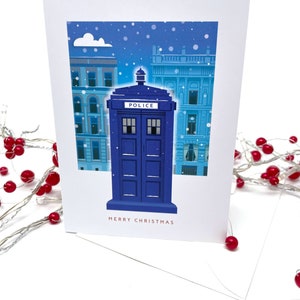 May include: A white Christmas card featuring a blue police box with the word "POLICE" on top, set against a snowy cityscape. The words "MERRY CHRISTMAS" are printed in red at the bottom.