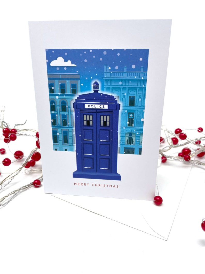 May include: A white Christmas card featuring a blue police box with the word "POLICE" on top, set against a snowy cityscape. The words "MERRY CHRISTMAS" are printed in red at the bottom.