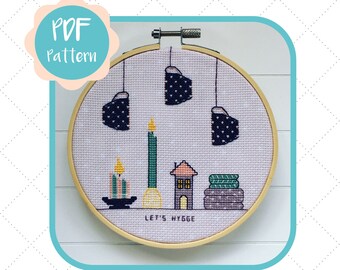 May include: A cross-stitch pattern featuring a cozy scene with a house, candles, and the text "Let's Hygge". The pattern is displayed in a wooden embroidery hoop.