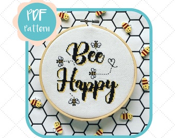 Può includere: Un cerchio da ricamo bianco con le parole "Bee Happy" ricamate in filo nero e giallo. Il cerchio è circondato da piccole figure di api gialle e nere su uno sfondo di piastrelle bianche.