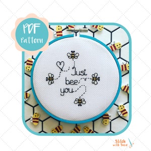 May include: A white embroidery hoop with a blue border, featuring a cross-stitch design of bees and the text "Just bee you". The hoop is surrounded by small wooden bee figurines on a white tiled background.