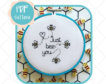 Può includere: Un cerchio da ricamo bianco con un bordo blu, che presenta un disegno a punto croce di api e il testo "Just bee you". Il cerchio è circondato da piccole figurine di api in legno su uno sfondo di piastrelle bianche.