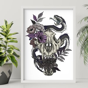 May include: A framed art print featuring a detailed illustration of a skull entwined with a snake and adorned with flowers. The artwork uses a palette of black, gray, purple, and white, creating a striking contrast. The print is displayed on a white surface.