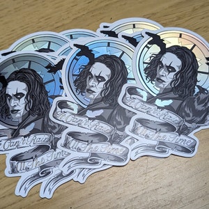 May include: Five silver holographic stickers featuring a black and white illustration of a man with long black hair and a black crow. The man is wearing a black coat and has a dark makeup look. The text on the sticker reads "It Can't Rain All The Time".