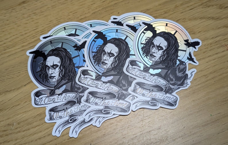 May include: Five silver holographic stickers featuring a black and white illustration of a man with long black hair and a black crow. The man is wearing a black coat and has a dark makeup look. The text on the sticker reads "It Can't Rain All The Time".