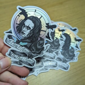May include: A stack of iridescent stickers featuring a black and white illustration of a man with long black hair and a black crow. The text on the sticker reads "It Can't Rain All The Time".