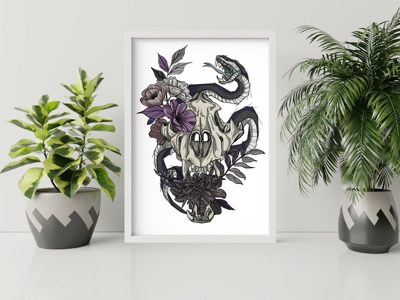 May include: A framed art print featuring a detailed illustration of a skull entwined with a snake and adorned with flowers. The artwork uses a palette of black, gray, purple, and white, creating a striking contrast. The print is displayed on a white surface.