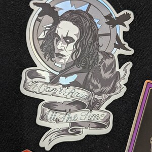 May include: A black and white sticker featuring a portrait of a man with long black hair and a black crow perched on his shoulder. The sticker has a circular design with a quote that reads "It Can't Rain All The Time".