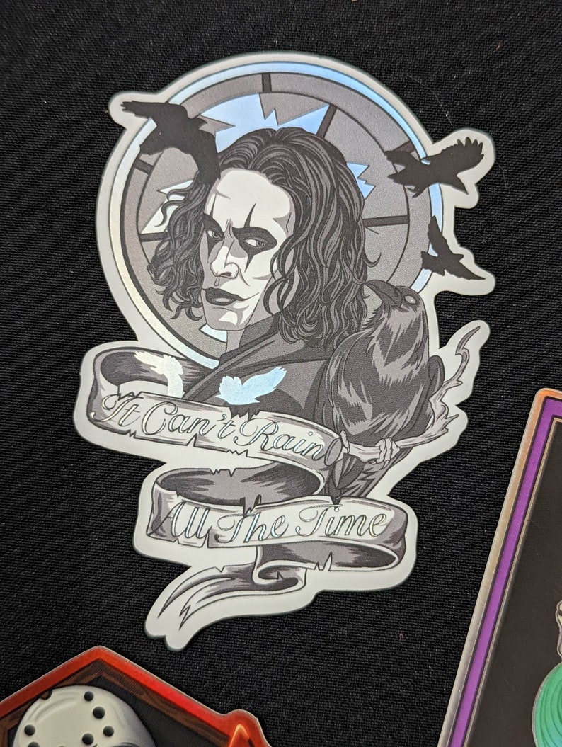 May include: A black and white sticker featuring a portrait of a man with long black hair and a black crow perched on his shoulder. The sticker has a circular design with a quote that reads "It Can't Rain All The Time".