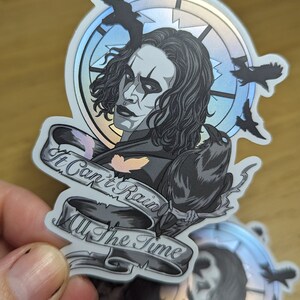 May include: A silver holographic sticker featuring a black and white illustration of a man with long black hair and a crow. The man is wearing a black shirt and has a dark makeup look. The sticker has a banner that reads "It Can't Rain All The Time".