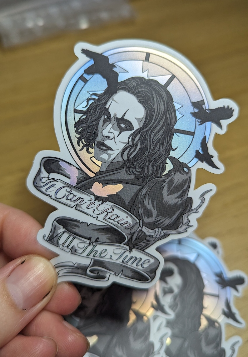 May include: A silver holographic sticker featuring a black and white illustration of a man with long black hair and a crow. The man is wearing a black shirt and has a dark makeup look. The sticker has a banner that reads "It Can't Rain All The Time".