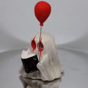 May include: A white ghost figurine holding a book with the word "IT" on the cover. The ghost has red eyes and is holding a red balloon.
