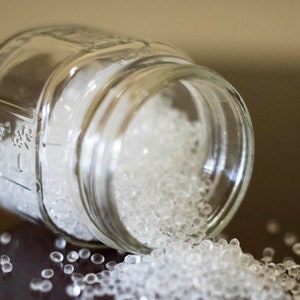 May include: A glass jar filled with clear silica gel beads, spilling out onto a dark brown surface. The jar is labeled with the text "Ball."