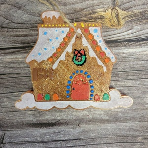 May include: A gingerbread house ornament with white icing, red and orange candy dots, and a green wreath with a red bow. The house has a red door and a white cloud base.