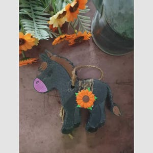 May include: A dark gray donkey ornament with a brown mane and tail, a pink nose, and a sunflower accent. The ornament has a jute rope for hanging and is decorated with a patterned saddle.