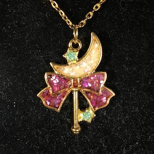 May include: A gold chain necklace with a pendant featuring a crescent moon, a pink bow, and a star. The pendant is decorated with glitter.