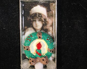 May include: A silver chain necklace with a rectangular pendant featuring a vintage photograph of a young girl wearing a white hat. The pendant has a green and red wreath with a white candle in the center.