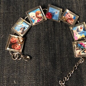 Pode incluir: Um bracelete de prata com múltiplos porta-retratos que apresentam personagens do filme Frozen. As molduras são decoradas com pequenos strass e um amuleto de flor vermelha.