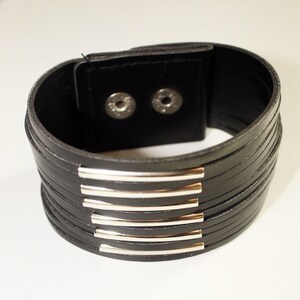 May include: A black leather bracelet with silver metal accents. The bracelet is made of multiple layers of leather and has a simple, modern design.