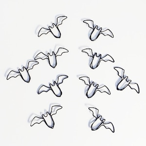 May include: A collection of nine small, silver-toned metal bat-shaped paper clips arranged in a scattered pattern. Each clip features a stylized bat silhouette with outstretched wings and a pointed ear design, creating a uniform look across the set.