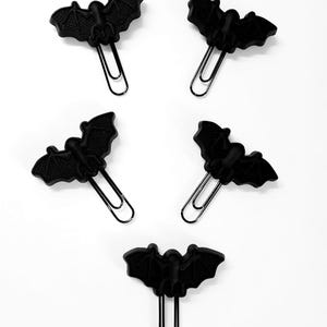 May include: Five black paper clips, each featuring a molded bat-shaped topper with detailed wing textures. The bats are matte black and attached to standard metal paper clips, arranged in a scattered pattern against a plain white background.