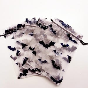Könnte beinhalten: Ein Set aus vier halbtransparenten weißen Organza-Beuteln mit Kordelzug, die mit einem sich wiederholenden Muster aus kleinen schwarzen Fledermäusen bedruckt sind. Jeder rechteckige Beutel hat oben eine dünne schwarze Kordel.