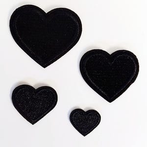 Könnte beinhalten: Vier schwarze, herzförmige Stickaufnäher in verschiedenen Größen, die in einer Gruppe angeordnet sind. Jeder Aufnäher verfügt über eine einfarbig schwarze, bestickte Oberfläche mit einem passenden schwarzen Rand, was ein einheitliches Bild ergibt.