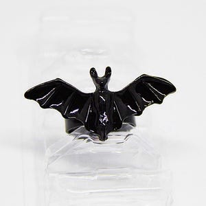 Può includere: Un anello nero a forma di pipistrello con le ali spiegate. L'anello è realizzato in materiale lucido e si staglia su uno sfondo bianco. È un accessorio a tema gotico.