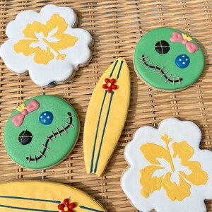 May include: Five decorated sugar cookies, including two green cookies with stitched faces and pink bows, a yellow surfboard cookie, and two white cookies with yellow hibiscus flowers.