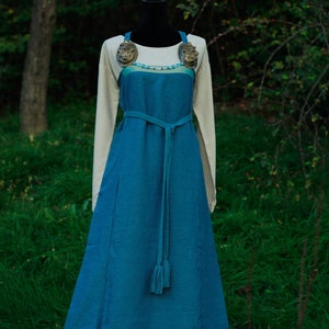 May include: A long, flowing blue dress with a white under layer and a decorative belt. The dress has a tassel at the end of the belt and two decorative metal pieces on the shoulders.