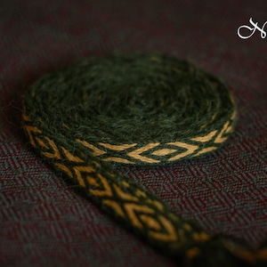 May include: A green and gold woven braid with a diamond pattern. The braid is rolled up and lying on a dark green and burgundy fabric.