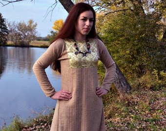 May include: A long, brown, medieval-style dress with a gold embroidered neckline and a beaded necklace. The dress is worn by a person standing in a grassy area with a lake in the background.