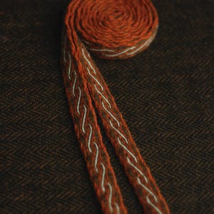 May include: A coiled woven band featuring a deep orange and brown base with a repeating light blue wave-like pattern running along the center. The textile strip has a textured appearance and ends in loose, frayed yarn strands at the bottom edges.