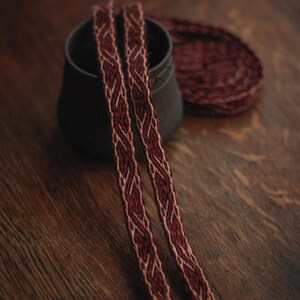 May include: A long, narrow woven band featuring a complex, repeating interlaced pattern in deep burgundy and soft beige yarn. The textile strip is draped over a dark, matte-finished ceramic vessel, with a coiled section visible in the background.