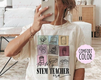 May include: White t-shirt with a graphic design of colorful squares featuring STEM-related icons, including a beaker, a laptop, a calculator, gears, a microscope, pencils, a coffee cup, safety goggles, and building blocks. The text "In my STEM Teacher era" is printed below the squares.