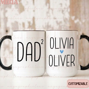 May include: Two white ceramic mugs with black handles. The mug on the left says "Dad^2" and the mug on the right says "Olivia Oliver" with a blue heart between the names. The mugs are customizable.