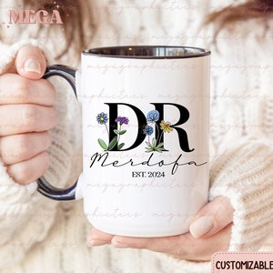 May include: White ceramic mug with a black rim and handle. The mug features a black and white design with the letters "DR" and the text "Merdofa EST. 2024". The letters are decorated with colourful flowers.
