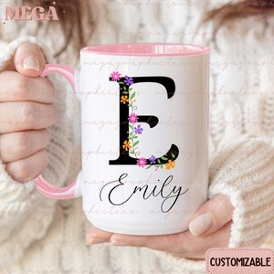 Pode incluir: Caneca de cerâmica branca com borda e alça rosa. A caneca apresenta uma letra "E" preta cercada por flores coloridas e o nome "Emily" escrito em itálico preto.