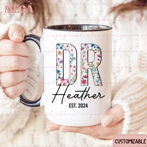 May include: White ceramic mug with a black rim and handle. The mug features a floral design with the letters "DR" in black outlined in white. Below the letters is the text "Heather EST. 2024".