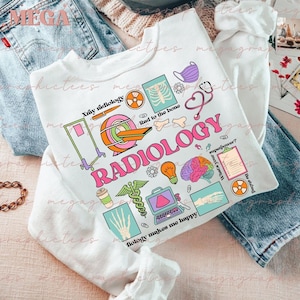 Könnte beinhalten: Ein weißes Sweatshirt mit einem bunten Grafikdesign, das das Wort "RADIOLOGY" und verschiedene medizinische Symbole zeigt, darunter ein Röntgenbild, ein Stethoskop, eine Glühbirne und ein Gehirn. Der Text "X-ray radiology", "Rad to the bone" und "Radiology makes me happy" ist ebenfalls im Design enthalten.