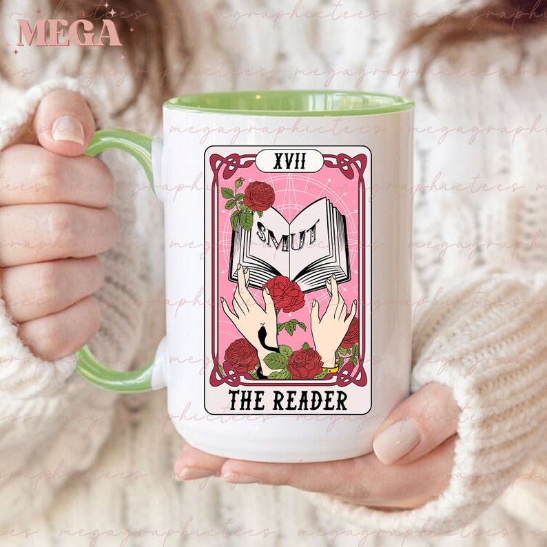 May include: A white ceramic mug with a green rim features a pink tarot card design. The card depicts a pair of hands holding open a book with the word "SMUT" printed on the pages. The card is labeled "XVIII THE READER".