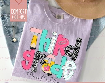 May include: A light purple t-shirt with the words "Third Grade" in a colorful, playful font. The design includes a pencil, apple, and other school-themed elements. The text "Mrs. Miller" is below the main text. The shirt is part of the "Comfort Colors" collection.
