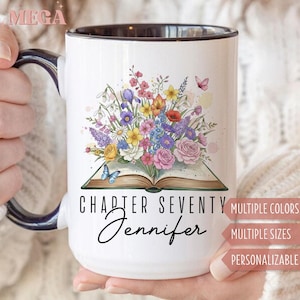 May include: White ceramic mug with a black interior and handle. The mug features a colorful floral design above an open book, with the text "CHAPTER SEVENTY Jennifer". Butterflies and flowers are also present.